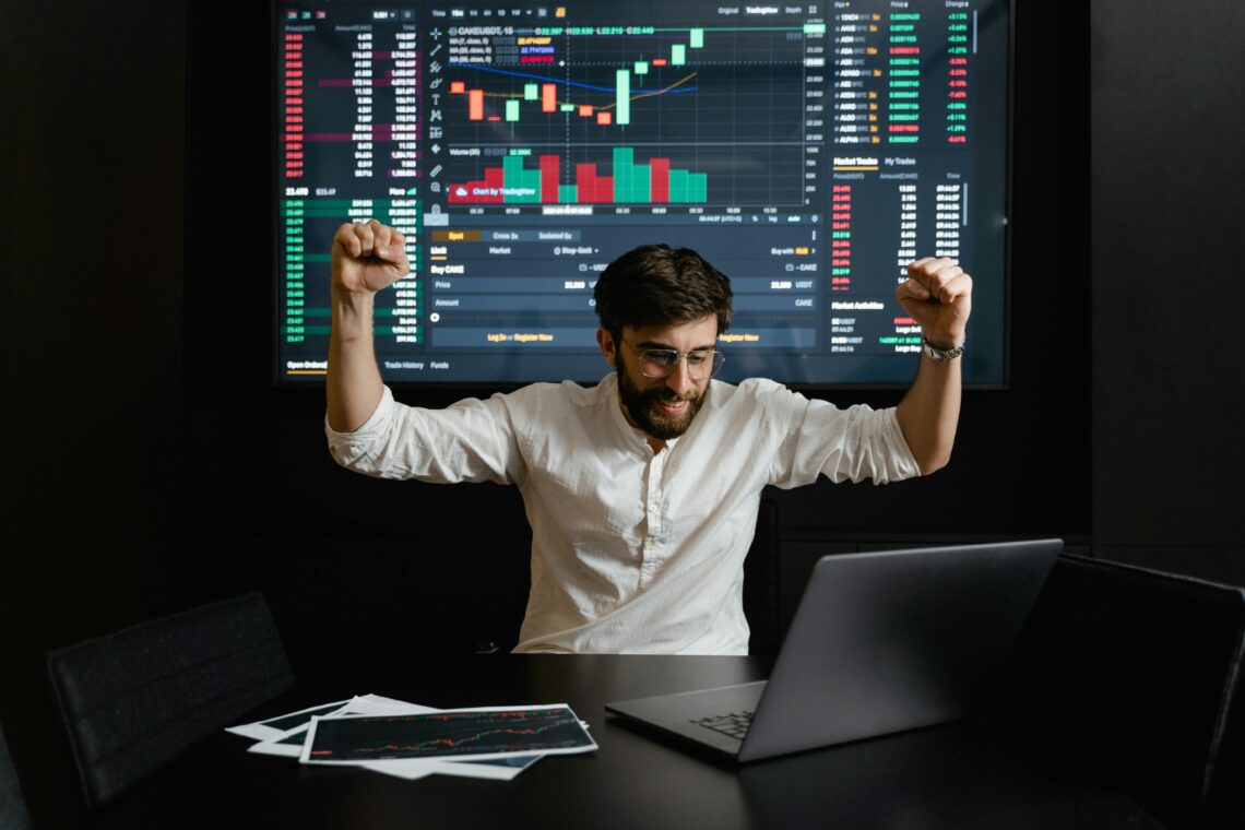 Excited trader celebrating in front of crypto trading charts on laptop and large screen, highlighting WLFI's performance as a new addition on MEXC.